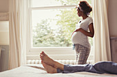 Pensive pregnant woman looking out bedroom window
