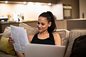 Smiling woman reading paperwork and using laptop on sofa