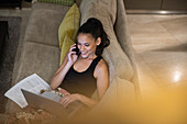 Woman talking on smart phone and using laptop on sofa