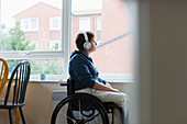 Thoughtful woman in wheelchair listening to music at window