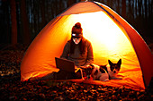 Woman with dog using laptop in glowing tent