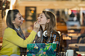 Woman applying lip gloss to friends lips in cafe window