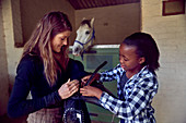 Woman helping girl prepare saddle in horse stables