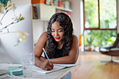 Woman writing in notebook at computer in home office