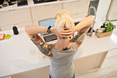 Young freelancer with tattoos working at laptop in kitchen