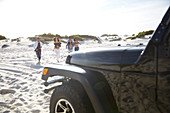 Young friends running on sunny beach toward jeep