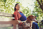Smiling pregnant woman with dog sitting on park bench