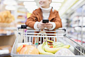 Woman with smart phone pushing shopping cart in supermarket