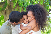 Parents kissing toddler daughter under tree