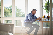 Senior man reading book at dining table by window