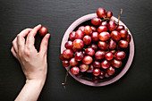Hand holding red grape near bowl of ripe red grapes placed on black surface
