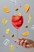 Crop female with glass of red alcohol cocktail garnished with orange slices and green olives above ground on gray background