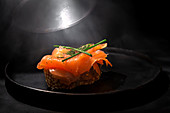 Smoked salmon and fennel herb on top of seeded bread on a dark plate and dark table