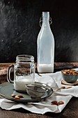Jar and bottle of vegan milk and bowl of almonds placed on rustic table near napkin and strainer