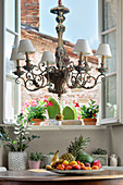 Ornate chandelier above plate of fruit on round antique table and potted plants in background