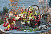 Kerzen in Töpfen mit Moos dekoriert mit Hagebutten, Korb mit Kleingeräten, Schnur und Stecketiketten, Zweige mit Hagebutten, Zieräpfeln und Schneebeeren