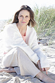 A long-haired woman sitting in the sand on the beach wearing a light jumper and trousers