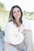 A long-haired woman sitting in the sand on the beach wearing a light jumper and trousers