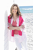 A long-haired woman on the beach wearing a long blouse with a pink jumper over her shoulders