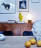 Blick über Esstisch auf Sideboard aus Teakholz mit Skulptur und Lampe, darüber Kunstwerke