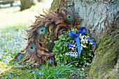 Boxwood wreath with a blue bow and flowers next to a wreath of peacock feathers
