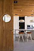 Wooden door with porthole window leading into modern kitchen with barstools at high table