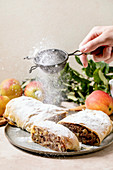 Homemade sliced classic apple strudel sprinkling by icing sugar from sieve in hand