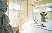 Woman in bathrobe fixing hair in sunny bathroom mirror