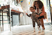 Happy mother helping baby daughter learn to walk at home