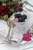 Still life with blackberries in glass bowls, next to a silver spoon and flowers