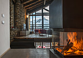 Fire in fireplace in front of modern lounge in open-plan interior with stone walls and wood-beamed ceiling