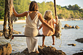Mother and daughter on large rope swing next to ocean, Thailand