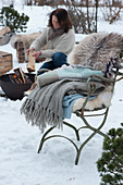 Woman puts firewood in the firepit, chair with blankets and fur