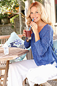 A blonde woman wearing a blue shirt and white trousers with a drink