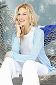 A blonde woman wearing a white top and a light blue cardigan sitting in a hammock