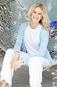 A blonde woman wearing a white top and a light blue cardigan sitting in a hammock