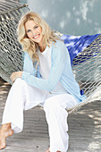 A blonde woman wearing a white top and a light blue cardigan sitting in a hammock