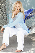 A blonde woman wearing a white top and a light blue cardigan sitting in a hammock