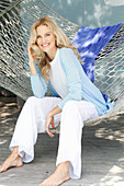 A blonde woman wearing a white top and a light blue cardigan sitting in a hammock