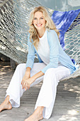 A blonde woman wearing a white top and a light blue cardigan sitting in a hammock