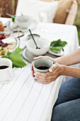 Frau hält Becher mit Kaffee am gedecktem Tisch auf der Terrasse