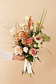 Hand holds bouquet with roses, calla lillies, chrysanthemums, carnations, amaranth, stonecrop, sea lavender and grass