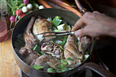 Hand holding tined fork stirs a copper pan of seared meat with leeks