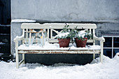 Laternen und Immergrünpflanzen auf weißer Holzbank im Schnee