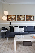 Row of photographs on wall above dark grey sofa, pendant lamps and coffee table on pale board floor in living room