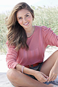 Langhaarige junge Frau in rosa Bluse und Shorts im Sand sitzend