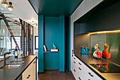 Illuminated fitted kitchen and kitchen island, turquoise wall with decorative shelf