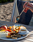 An outdoor table with a cheeseboard with feta, parmesan, crackers, pistachios, peaches and thyme, with honey being drizzled overtop