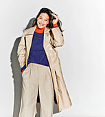 A dark-haired woman wearing an orange turtleneck, a jumper, a beige coat and trousers