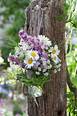 A small bouquet of lilacs, daisies, and caraway in a jar with a grass wreath hung on a post